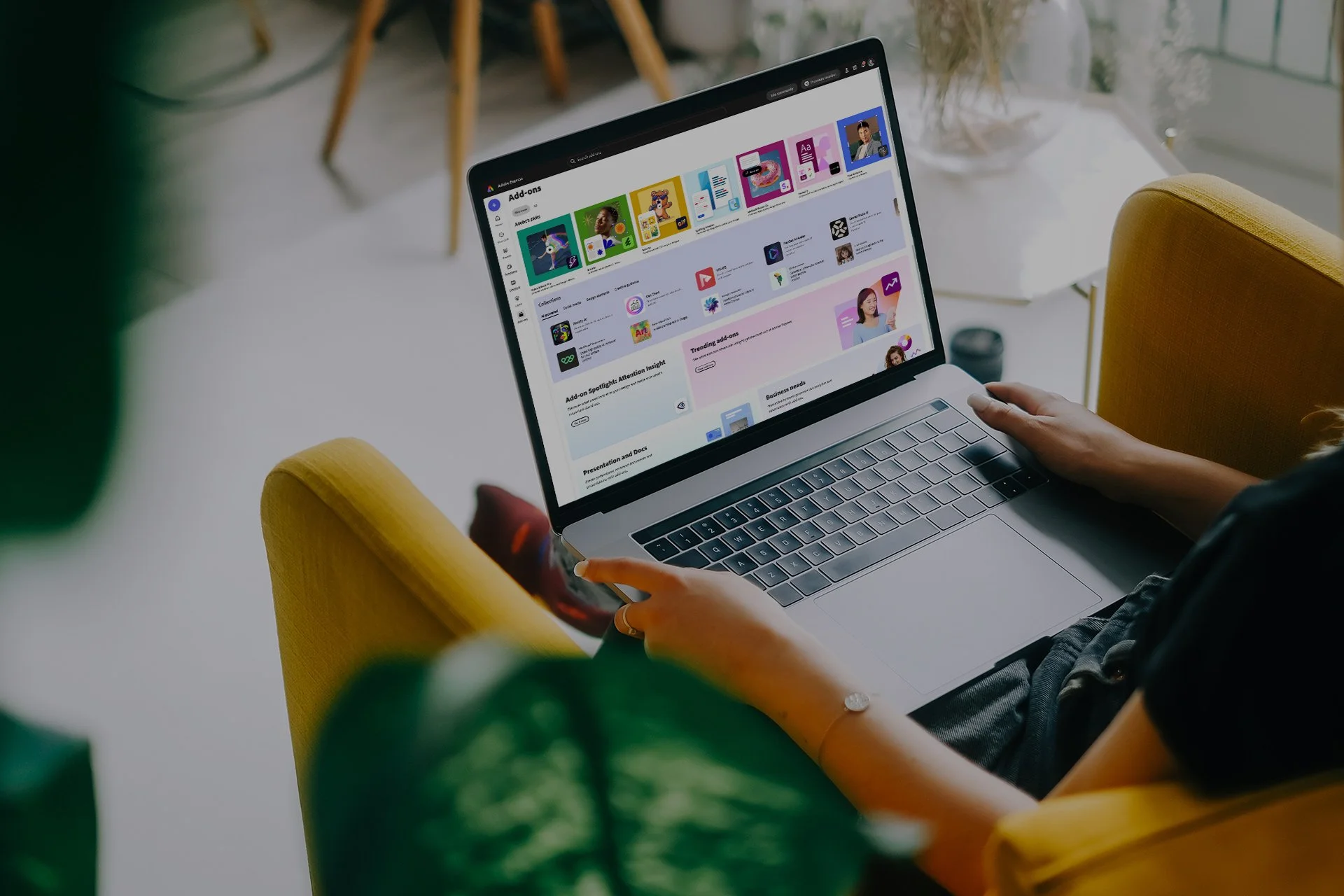Person using a laptop on a yellow armchair, browsing the Adobe Express Add-ons marketplace.