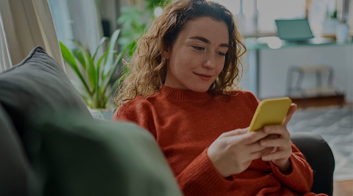 Woman relaxing on a sofa at home, smiling while looking at a yellow smartphone — an everyday shopping moment framing Walmart's AI shopping experience.